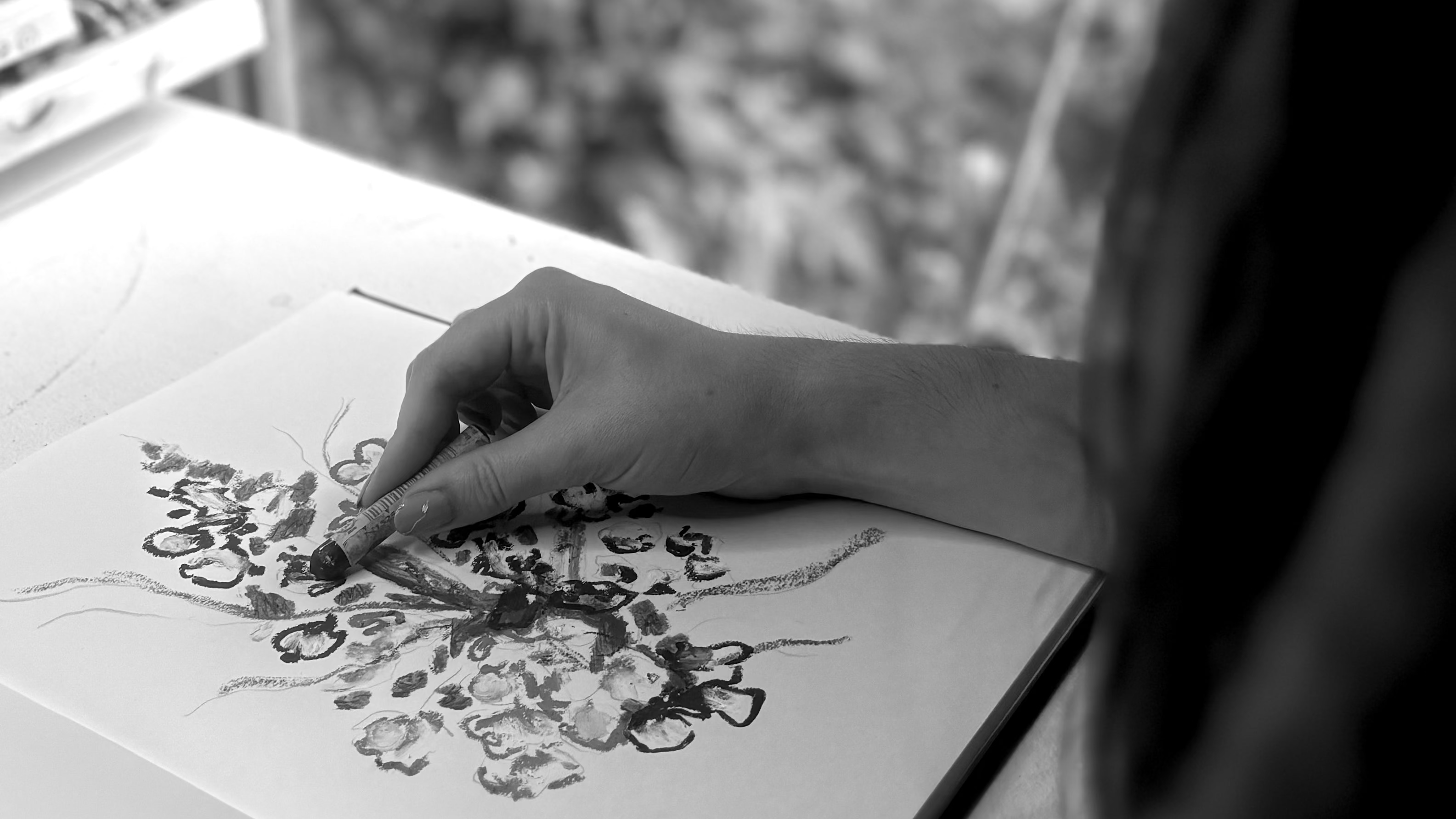 A black and white photograph of a person's hand drawing on a sheet of paper at a table, with a blurred window and foliage in the background.