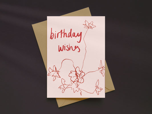 A square birthday card with a pale blush background and red handwritten-style text reading 'birthday wishes,' decorated with simple red line-drawn flowers, shown with a kraft envelope on a dark charcoal background.