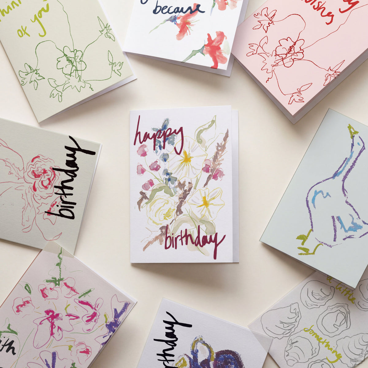 A set of colorful greeting cards arranged in a flat-lay circle on a light cream background. The central card is white with a watercolor floral design and the words 'happy birthday' in red and purple script.
