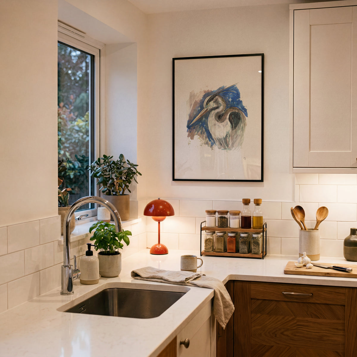 A framed abstract art print featuring a blue and white heron hangs on a warm beige wall in a modern kitchen. The kitchen has white subway tiles, a white marble countertop with a stainless steel sink, a red dome-shaped table lamp, and wooden cabinets.